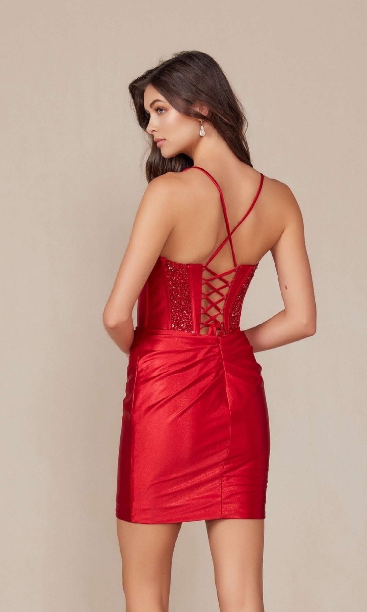 Woman in the Embellished-Corset Mini Homecoming Dress R947—a shiny, fitted red mini dress with lace-up back, thin straps, and sparkly embellishments—stands against a plain beige background, facing away from the camera.